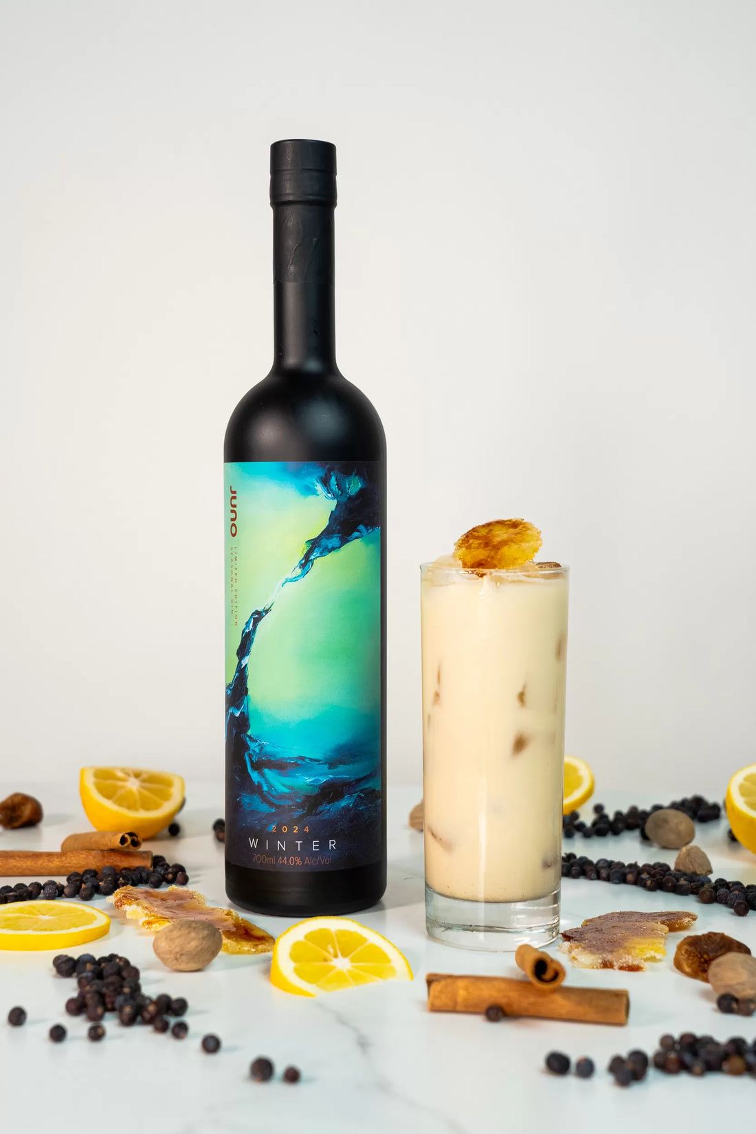 A Gin Brulee cocktail in a tall glass surrounded by spices and lemon next to a bottle of Juno Gin
