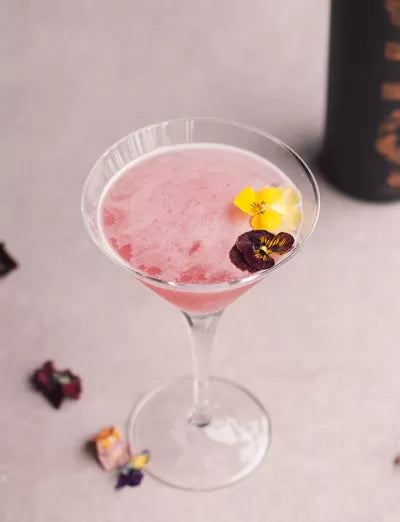 The Blushing Goddess cocktail in a martini glass, pink with a flower garnish
