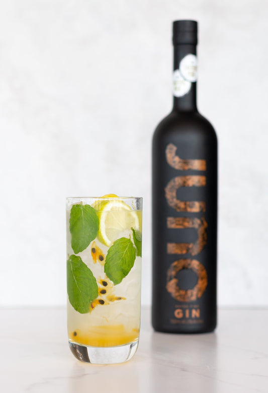 A Passionfruit Punch cocktail in a tall glass with mint next to a bottle of Juno Extra Fine Gin