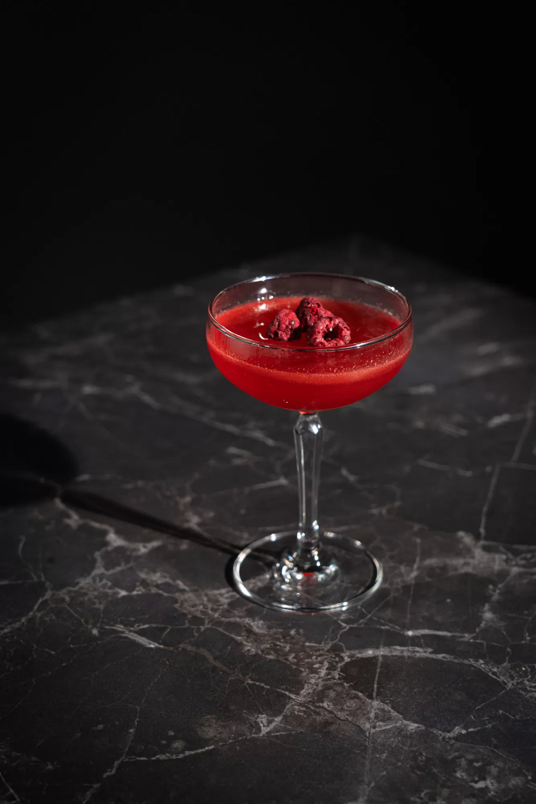 A bright red Raspberry Beret cocktail in a coupe glass with raspberries on top