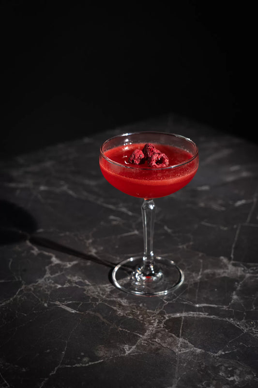 A bright red Raspberry Beret cocktail in a coupe glass with raspberries on top