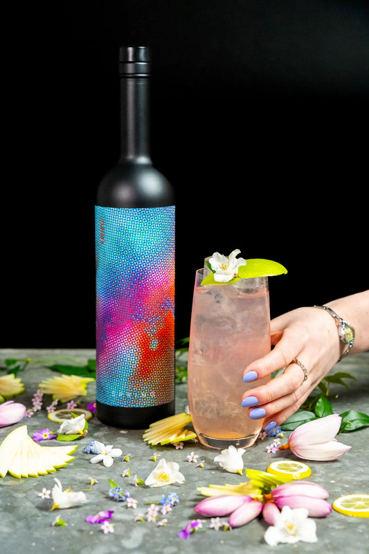 A light pink Frolic cocktail with an apple blossom garnish and a bottle of Juno Spring 2025 Seasonal Gin