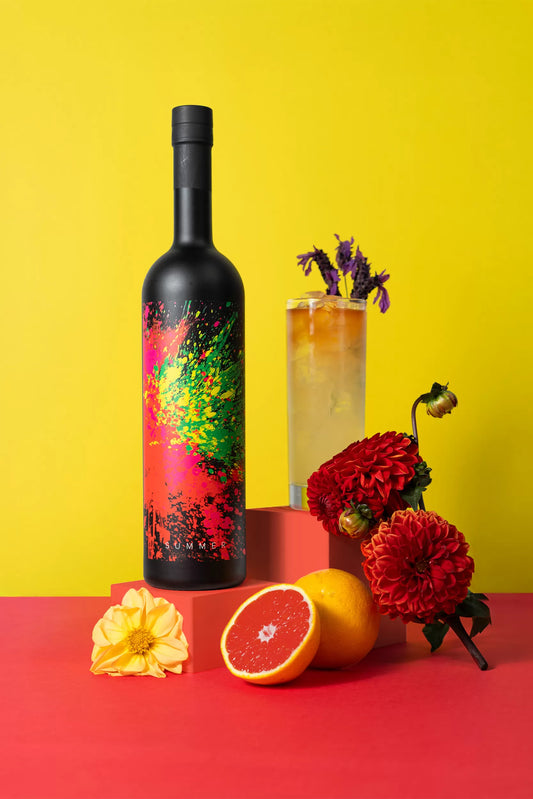 A Bitter Beach cocktail in a tall glass next to a bottle of Juno Summer 2025 Seasonal Gin and some citrus fruit