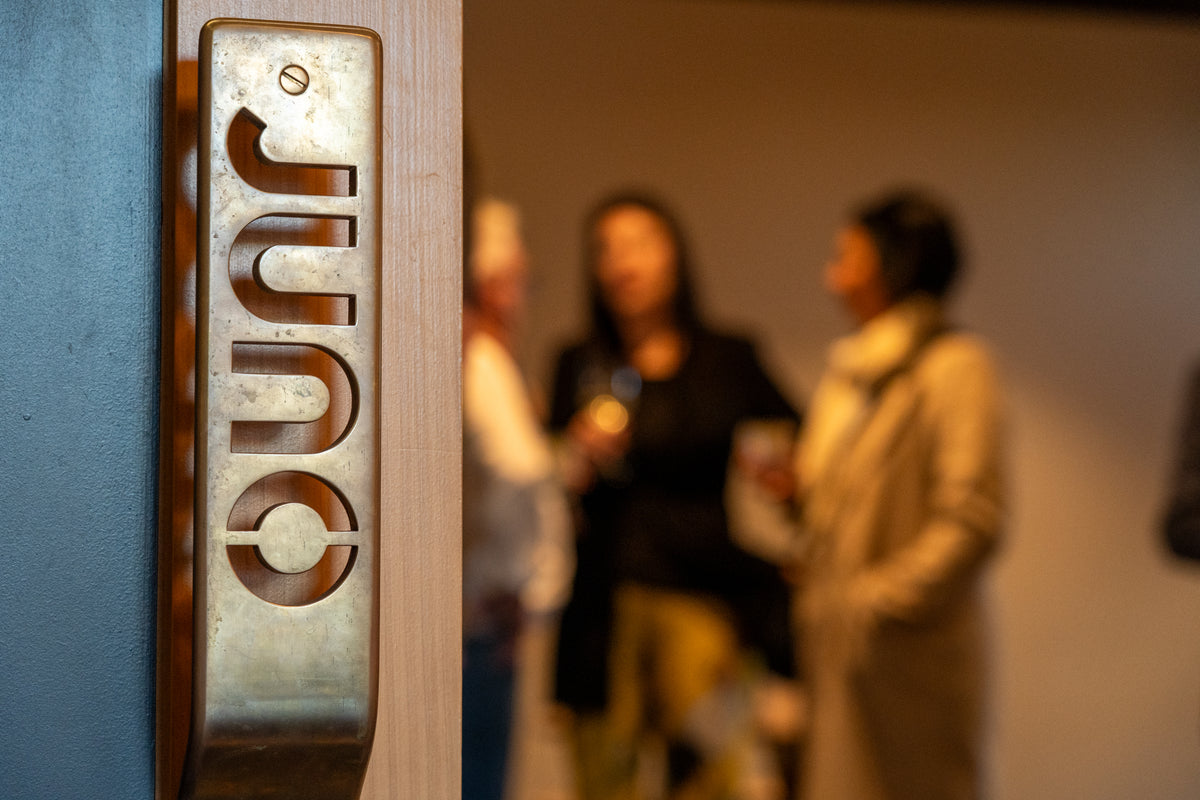 A peek through the door of the Tasting Room. A gold handle with JUNO written on it in foreground, people enjoying a cocktail in background. 