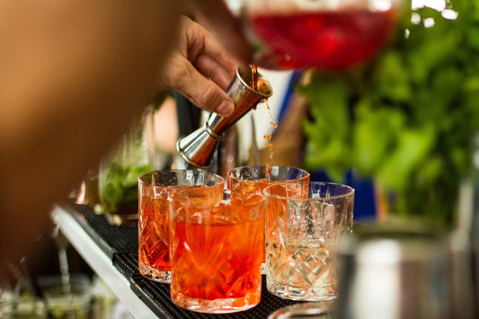 A close-up action shot of a bartender pouring a vibrant red-orange cocktail from a glass pitcher into a metal jigger, with several textured glasses filled with the drink on a bar counter. The background is a soft-focus bar setting with green foliage.